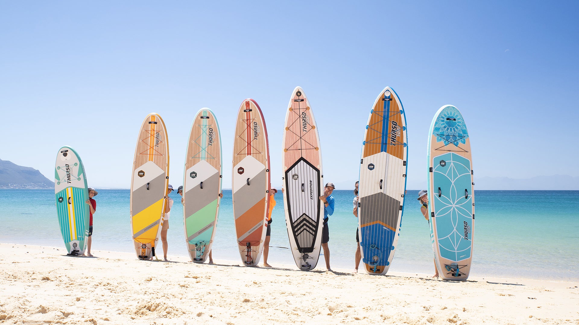 Eine Vielzahl bunter Paddleboards ist an einem sonnigen Strand ausgestellt und spiegelt eine Reihe von Designs für verschiedene Wasseraktivitäten wider.