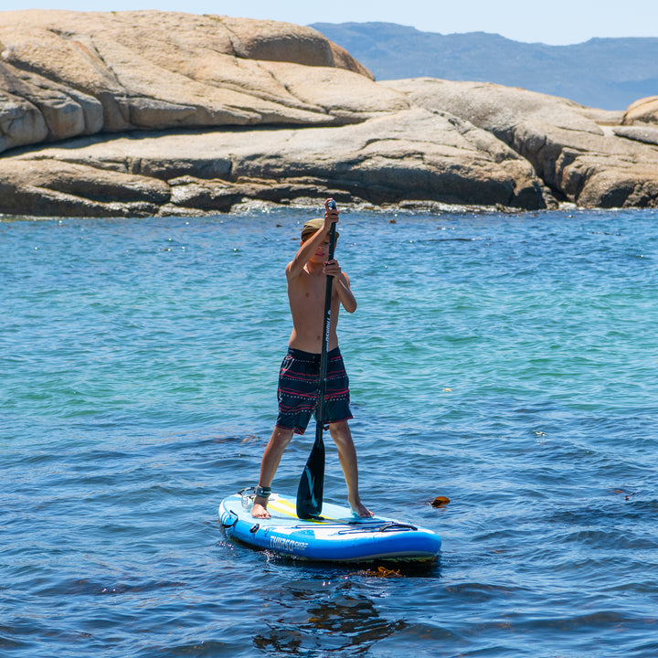 A young paddler standing on a bright azure Thurso Surf Prodigy junior paddle board, navigating clear ocean waters near a rocky shoreline