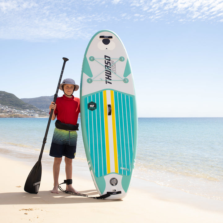 A young paddler standing on a sandy beach next to an upright Emerald green Thurso Surf Prodigy paddle board, holding a black paddle against a blue ocean background.