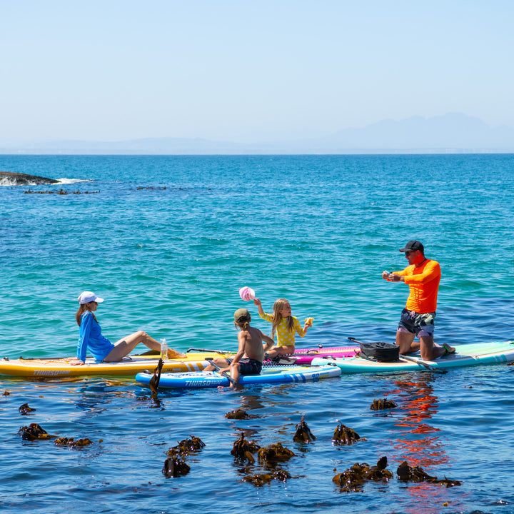 A group of paddlers on the ocean, including a child on a Magenta Thurso Surf Prodigy board, enjoying a sunny day.