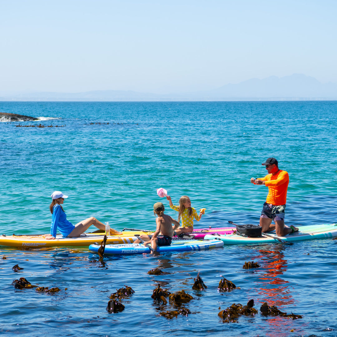 A group of paddlers on the ocean, including a child on a Magenta Thurso Surf Prodigy board, enjoying a sunny day.