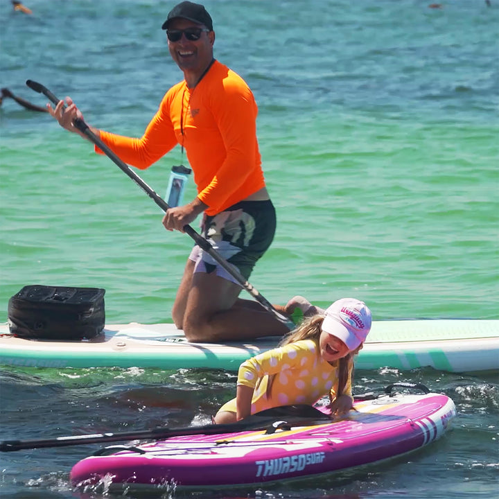 A happy child jumping onto a Magenta Thurso Surf Prodigy paddle board in the ocean while an adult on a green board looks on and smiles.