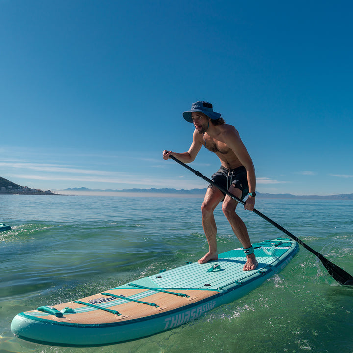 A person stand-up paddleboarding on a calm, clear day. They are using a turquoise paddle board, with scenic mountains visible in the background.