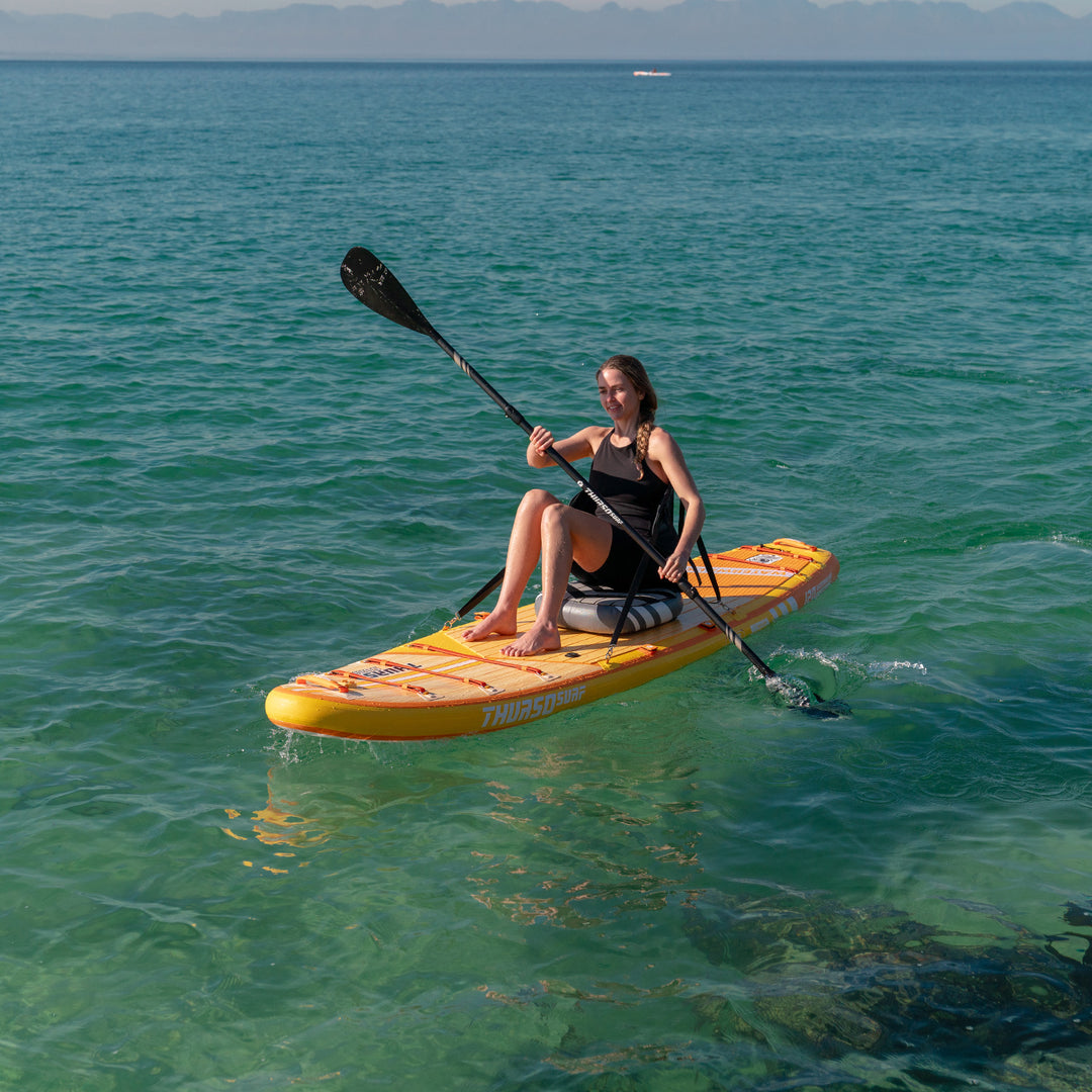 A woman paddleboarding on a clear blue ocean with mountains in the background, using a Thurso Surf Waterwalker 126 Tangerine paddleboard.